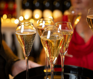 Several champagne flutes filled with sparkling wine are arranged on a black tray, with a person in a red dress holding one flute in a warmly lit bar setting.